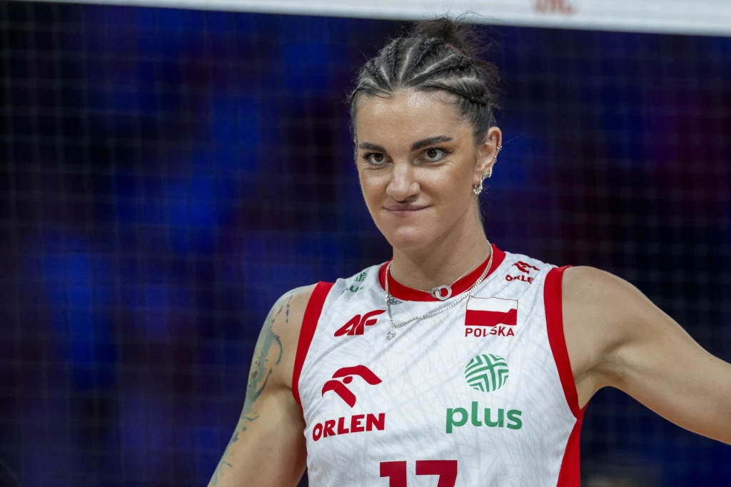 Siatkarka w koszulce reprezentacji Polski ze skupionym wyrazem twarzy, widoczne są warkoczyki oraz sportowa atmosfera na hali, tło rozmyte z siatką.