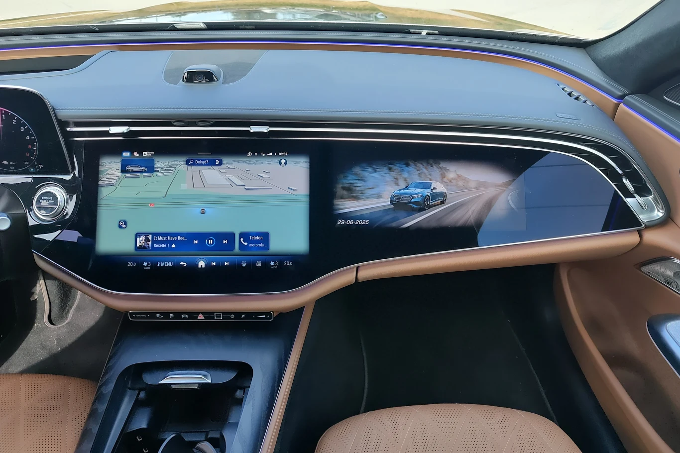 Kokpit nowoczesnego samochodu z rozbudowanym, szerokim panelem multimedialnym obejmującym ekran nawigacji i ekran z wizualizacją auta, fotele z jasnej skóry, elementy wykończenia w kolorze brązowym.