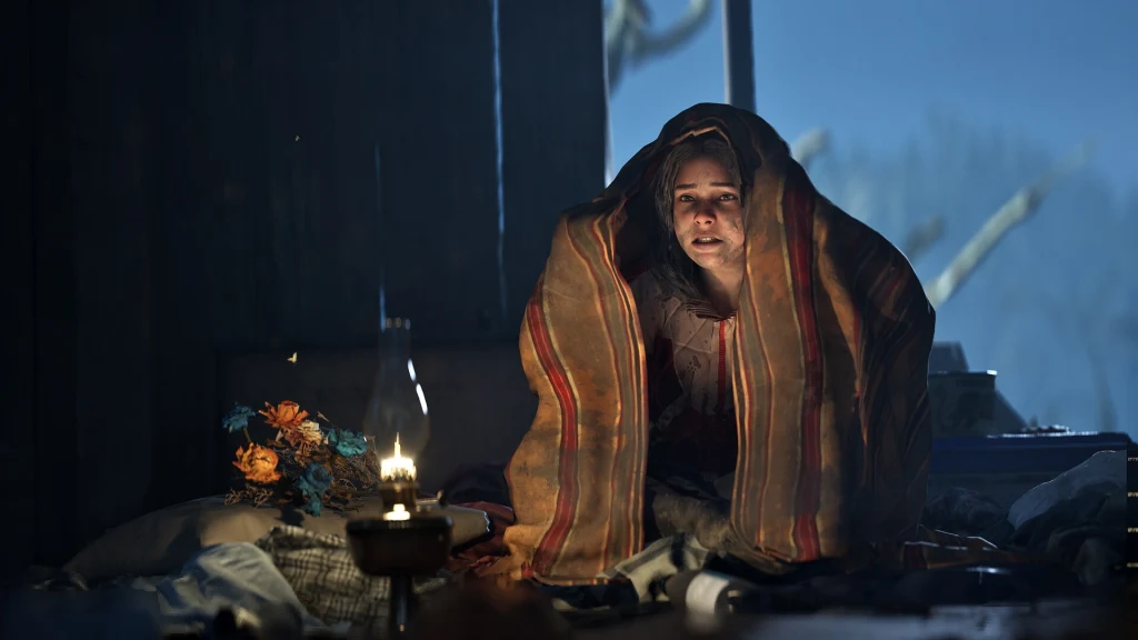 Hell is Us Młoda kobieta otulona kocem siedzi w ciemnym pomieszczeniu, oświetlona małą lampą naftową, wokół rozrzucone są przedmioty, a obok niej leży bukiet sztucznych kwiatów.