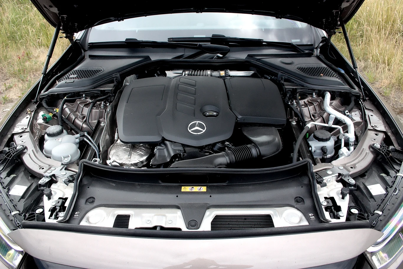 Komora silnika samochodu marki Mercedes, centralnie umieszczony silnik z wyraźnie widocznym logo producenta, otoczenie części mechanicznych i drobnych podzespołów, całość utrzymana w czystości.