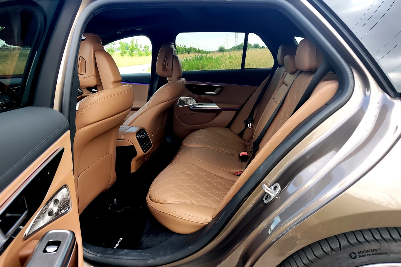 Brązowe skórzane fotele samochodu, przestronna tylna kanapa, eleganckie wykończenia drzwi oraz widok przez uchyloną szybę na zielony krajobraz.