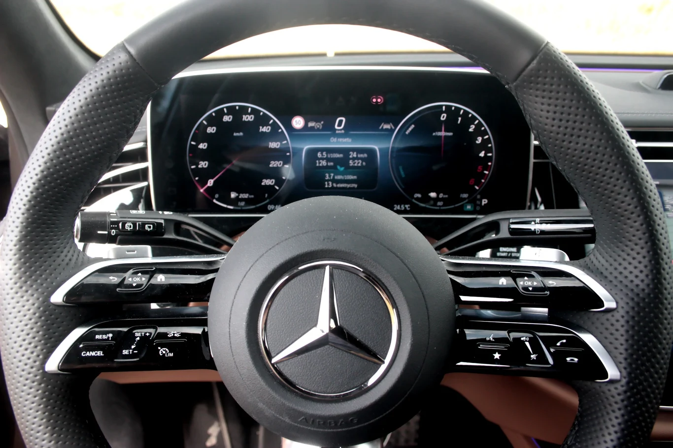 Nowoczesna kierownica samochodu marki Mercedes z wyraźnie widocznym logo pośrodku, ekran cyfrowych zegarów z tyłu ukazujący parametry jazdy oraz estetyczne wykończenie i przyciski sterujące na kierownicy.