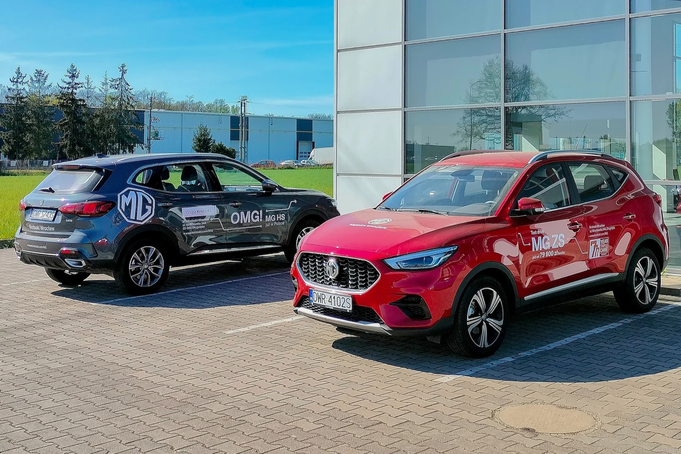 Porównaliśmy MG ze Skodą i Toyotą. Ile kosztuje serwis chińskich aut? Dwa nowoczesne samochody marki MG zaparkowane na wybrukowanym parkingu obok przeszklonego budynku, czerwony MG ZS na pierwszym planie po prawej i ciemny MG HS po lewej, wyraźne oznaczenia pojazdów promocyjnych.