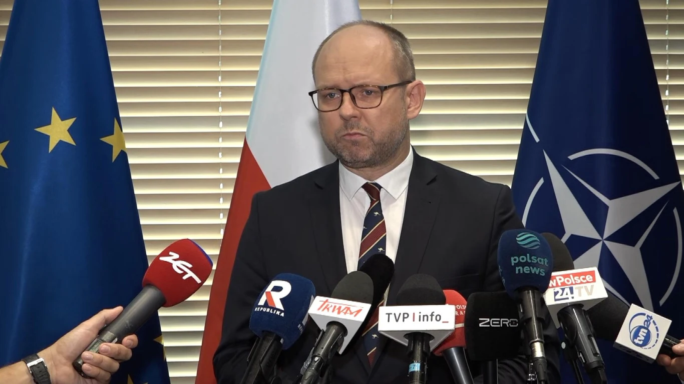 Marcin Przydacz o szczegółach wizyty prezydenta w Waszyngtonie Mężczyzna w garniturze i okularach stoi za pulpitem podczas konferencji prasowej, otoczony mikrofonami wielu różnych mediów, w tle widoczne są flagi Unii Europejskiej, Polski oraz NATO.