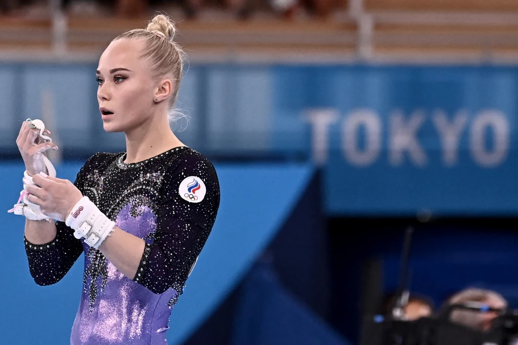 Angelina Mielnikowa podczas Igrzysk Olimpijskich w Tokio wywalczyła m.in. złoty medal Młoda gimnastyczka w sportowym stroju fioletowo-czarnym przygotowuje się do startu podczas zawodów, trzymając kawałek białej chusteczki, w tle widoczne elementy sali sportowej i napis TOKYO.