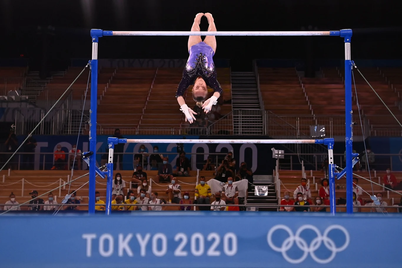 Gimnastyczka wykonująca skok na drążku podczas igrzysk olimpijskich w Tokio 2020, w tle widownia i oznaczenia olimpijskie.