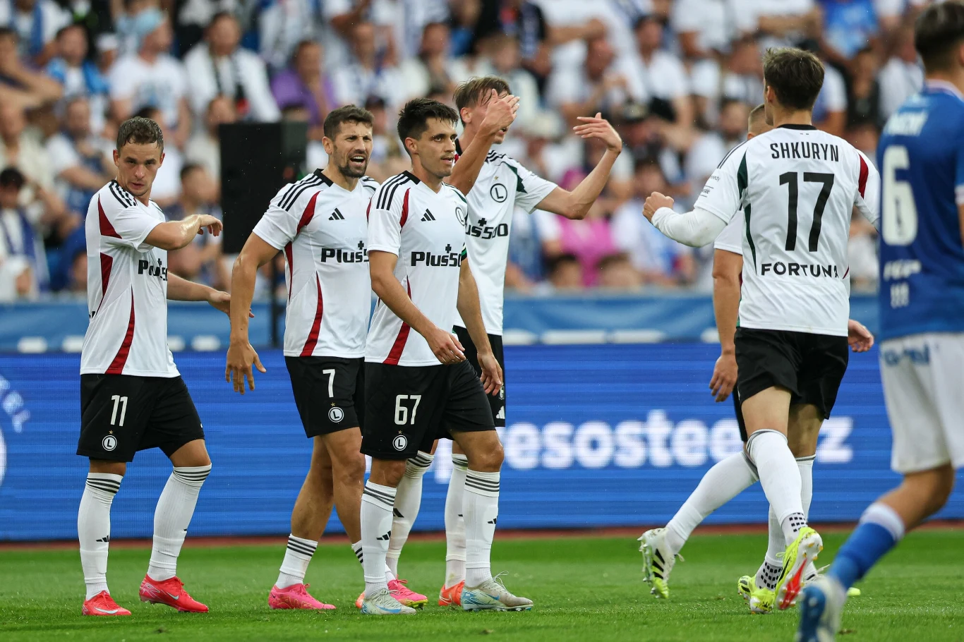 Piłkarze drużyny Legia Warszawa świętują zdobycie gola na boisku, ubrani w klubowe stroje, otoczeni przez innych zawodników oraz kibiców widocznych na trybunach.