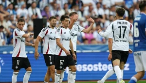 Piłkarze drużyny Legia Warszawa świętują zdobycie gola na boisku, ubrani w klubowe stroje, otoczeni przez innych zawodników oraz kibiców widocznych na trybunach.