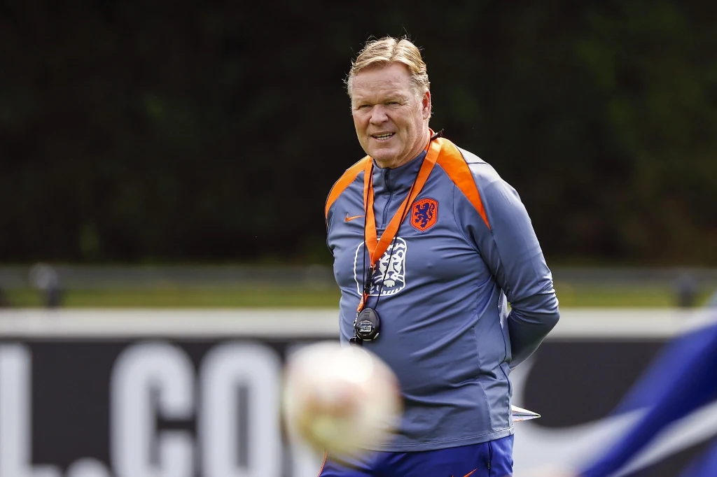 Trener piłkarski ubrany w sportową odzież z herbem reprezentacji Holandii stoi na boisku, z gwizdkiem na szyi, w tle rozmyte elementy boiska i piłka na pierwszym planie.