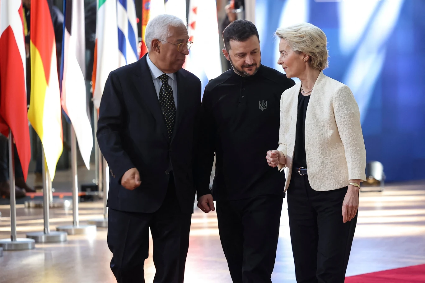 Trzech polityków idzie wspólnie, rozmawiając i wymieniając się spostrzeżeniami, w tle widoczne są flagi krajów europejskich ustawione w rzędzie.