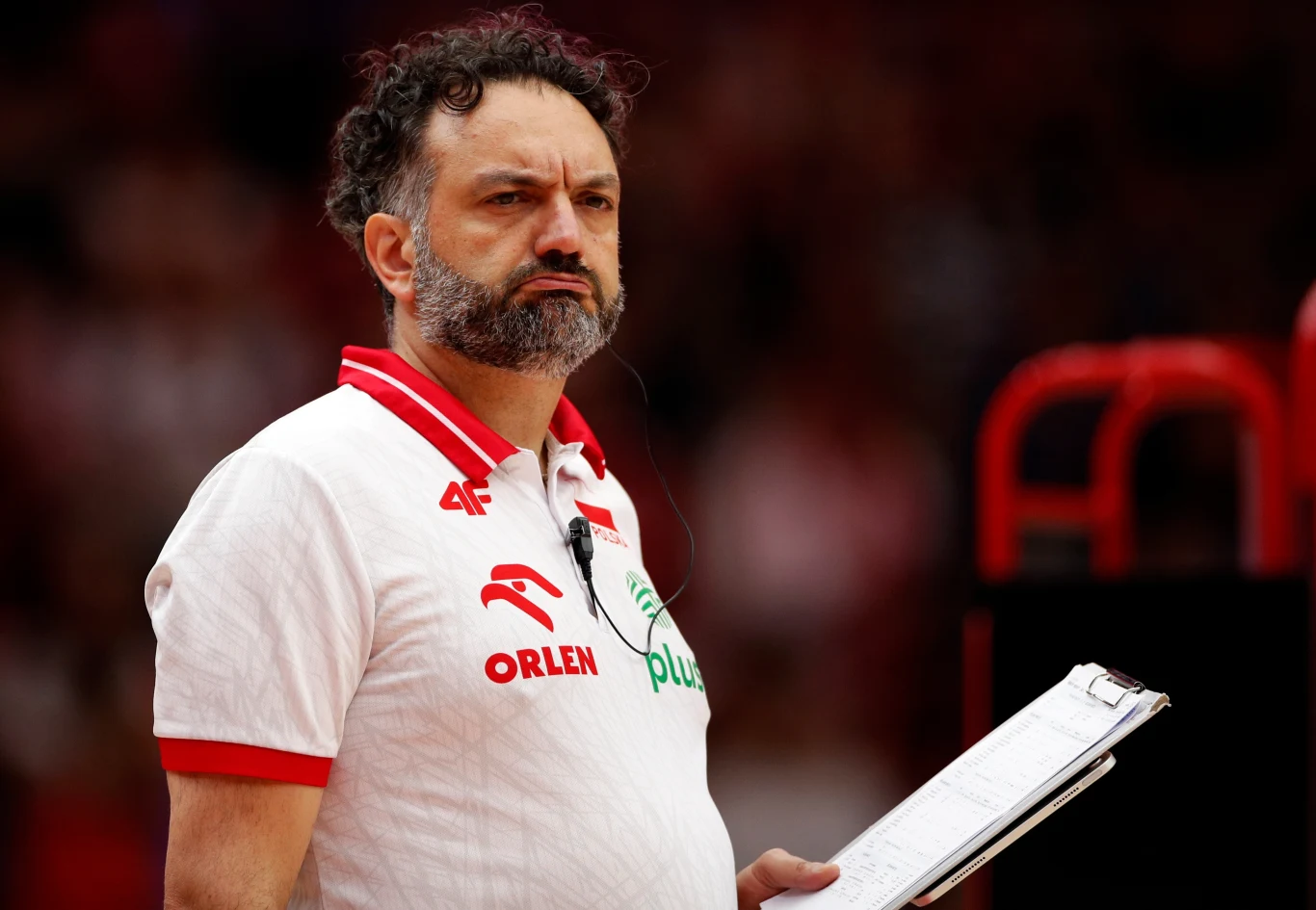 Mężczyzna z brodą w białej koszulce z logo sponsorów i napisem Orlen oraz Plus trzymający notatnik; koncentracja i powaga na twarzy sugerują rolę trenera podczas wydarzenia sportowego.