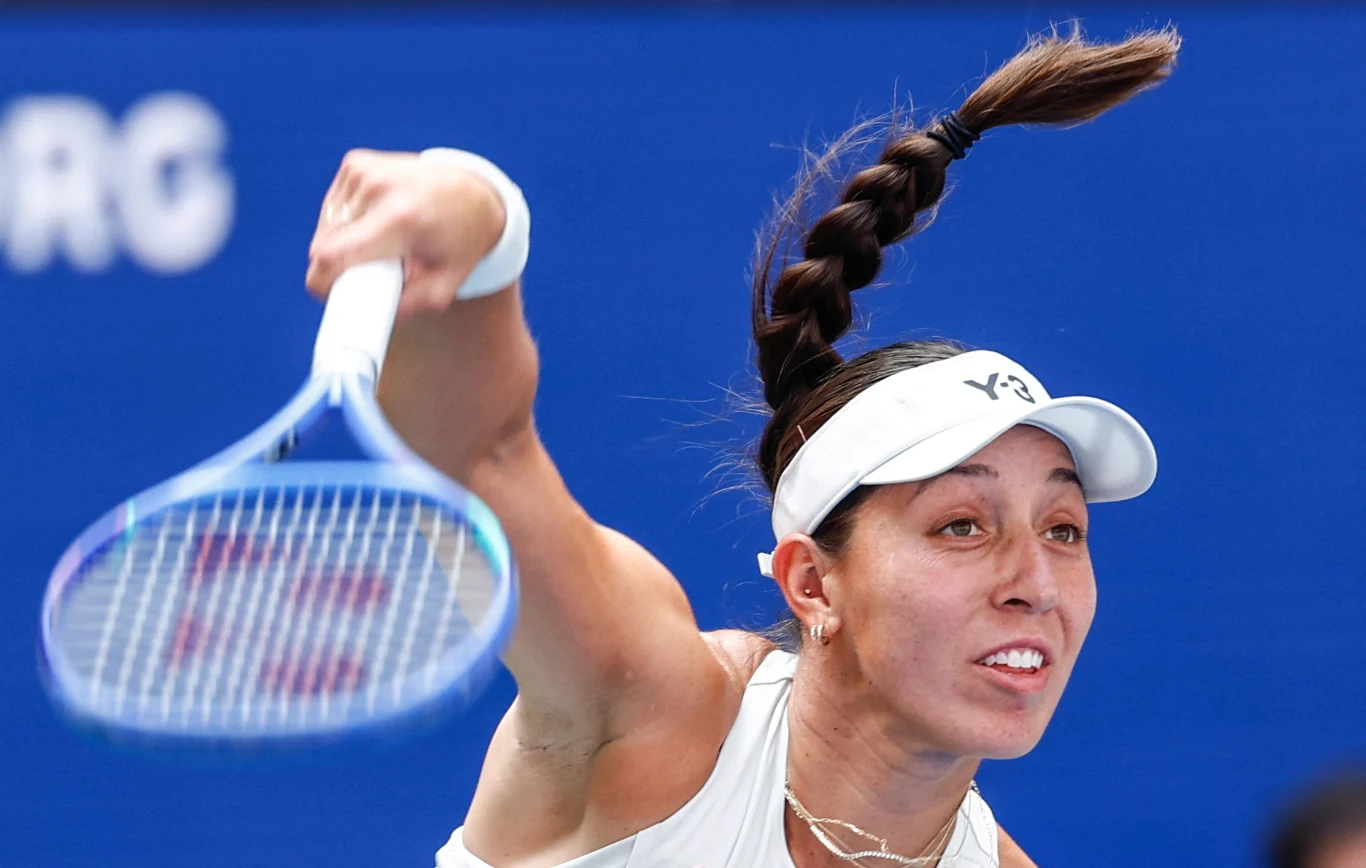 Jessica Pegula Zawodniczka wykonująca dynamiczny serwis podczas meczu tenisowego, skupiona twarz, napięta sylwetka i wyraźnie widoczna rakieta tenisowa, kontrastujące z niebieskim tłem kortu.