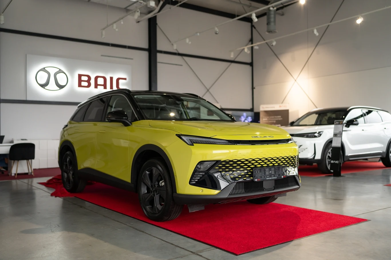 Nowoczesny, limonkowy samochód elektryczny marki BAIC eksponowany na czerwonym dywanie w jasnym, przestronnym salonie samochodowym, obok innego pojazdu elektrycznego i logo firmy na ścianie.