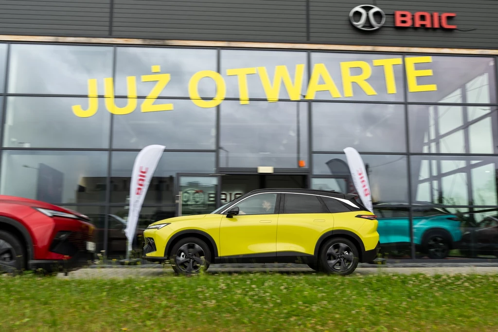 limonkowy samochód typu SUV zaparkowany przed nowoczesnym salonem samochodowym z wielkim napisem już otwarte oraz logo BAIC na fasadzie budynku, w tle widoczne są flagi reklamowe oraz inne auta