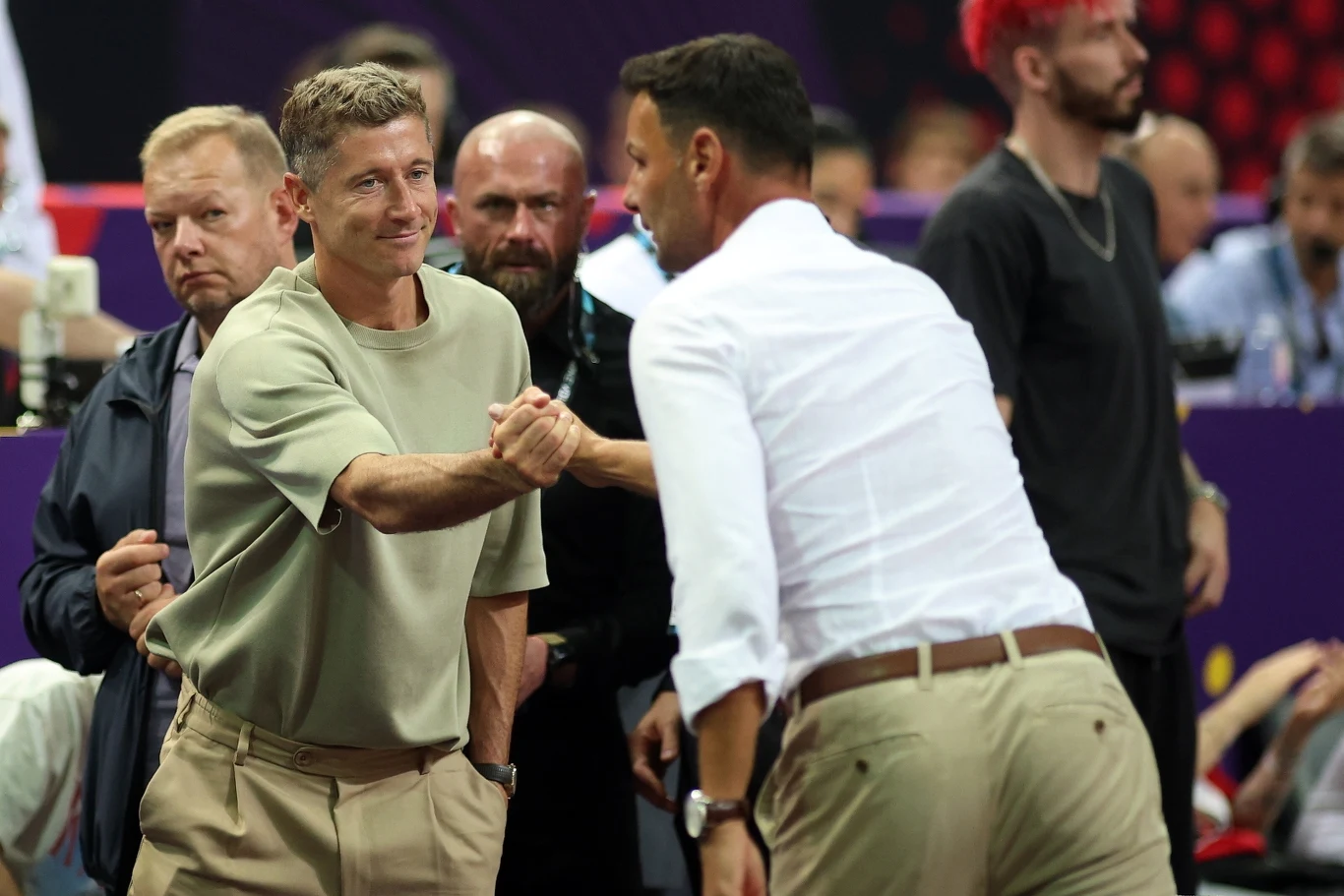 Igor Milicić przybił piątkę z Robertem Lewandowskim Dwóch mężczyzn w serdecznym uścisku dłoni w oficjalnej atmosferze, jeden ubrany w jasny t-shirt i beżowe spodnie, drugi w białą koszulę i beżowe spodnie, w tle kilku obserwujących mężczyzn.