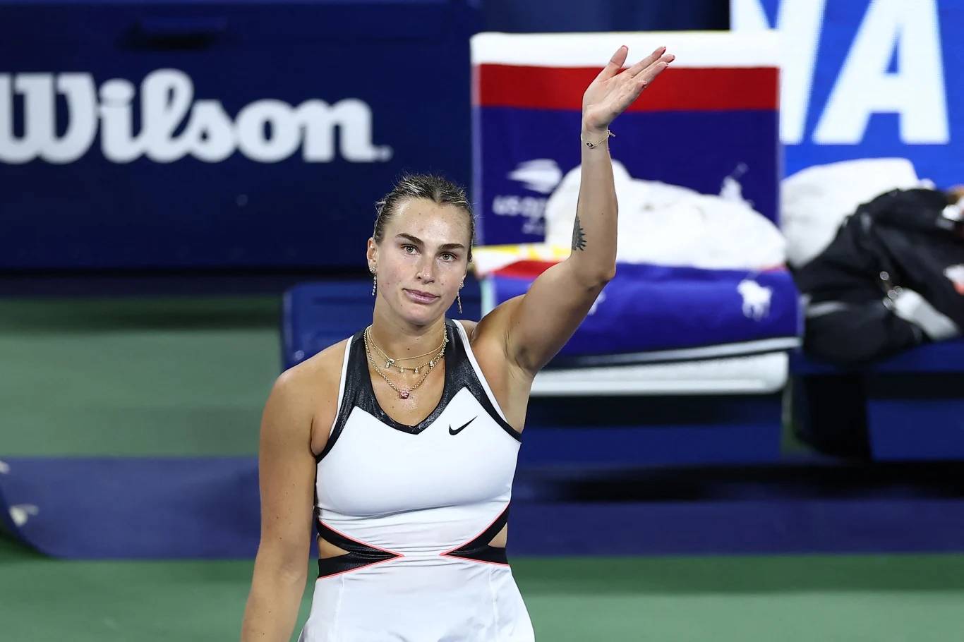 Aryna Sabalenka Zawodniczka tenisowa w stroju sportowym na korcie, unosi jedną rękę w geście pożegnania lub podziękowania, w tle widoczne ławki oraz logo sponsora.