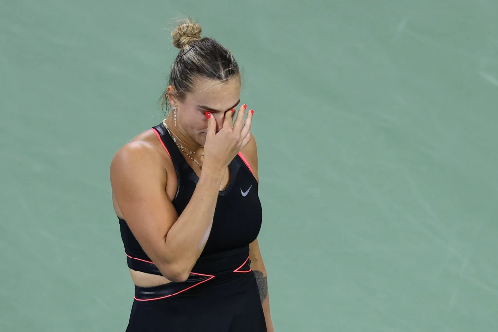 Aryna Sabalenka Sportowa zawodniczka w czarnym stroju na korcie, pochylająca głowę i zasłaniająca twarz dłonią, wyrażająca emocje po meczu.