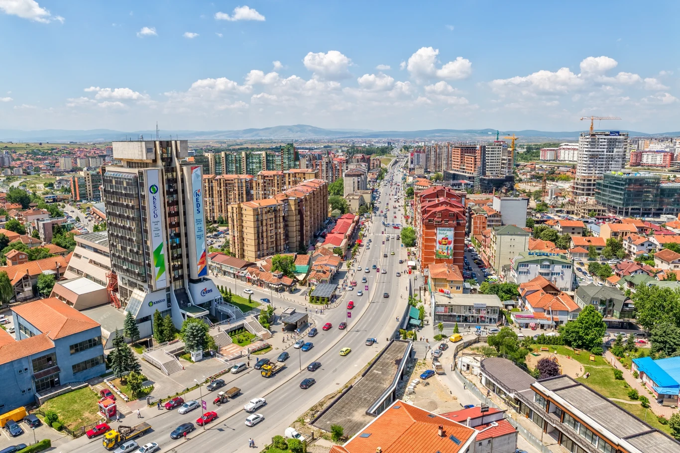 Kosowo, stolica miasta - Prisztina Ruchliwa arteria przechodząca przez centrum miasta otoczona nowoczesnymi wieżowcami, blokami mieszkalnymi oraz niską zabudową, widoczne liczne pojazdy na drodze, na horyzoncie widać góry i niebo z chmurami.