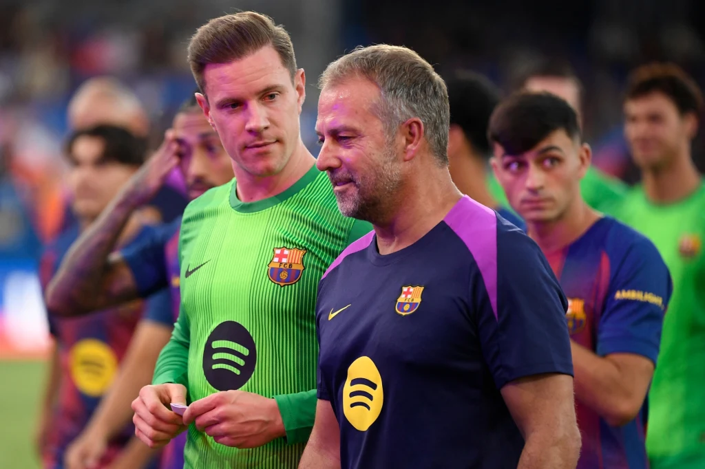 Piłkarz drużyny FC Barcelona ubrany na zielono oraz trener w granatowej koszulce z emblematem klubu rozmawiają na boisku, wokół nich stoją inni członkowie zespołu w klubowych strojach sportowych.