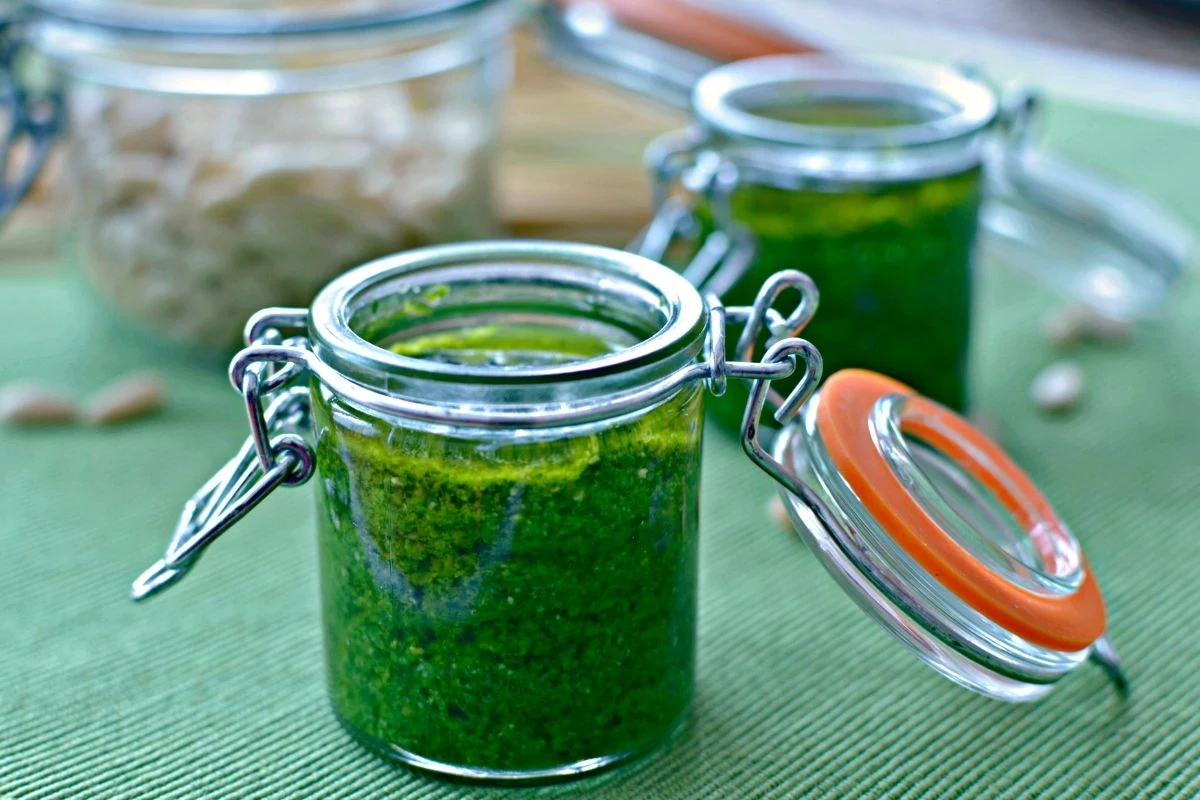 Słoik z zielonym sosem, najprawdopodobniej pesto, ustawiony na zielonej powierzchni, w tle drugi słoik z podobną zawartością oraz rozmyte nasiona słonecznika.