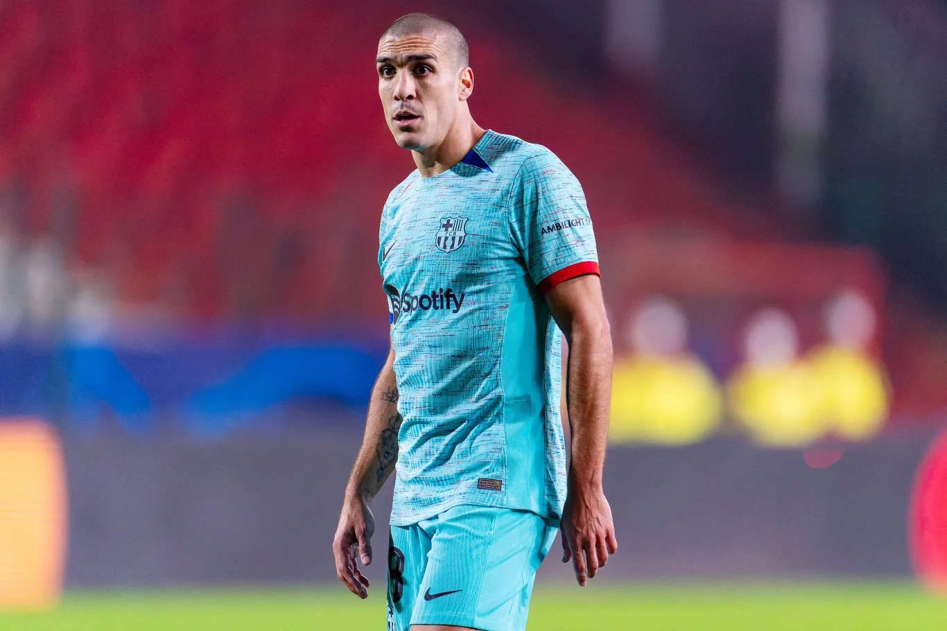 Oriol Romeu Piłkarz w jasnoniebieskiej koszulce z logo Spotify stoi na murawie stadionu, w tle rozmyte trybuny i żółte sylwetki ochroniarzy.