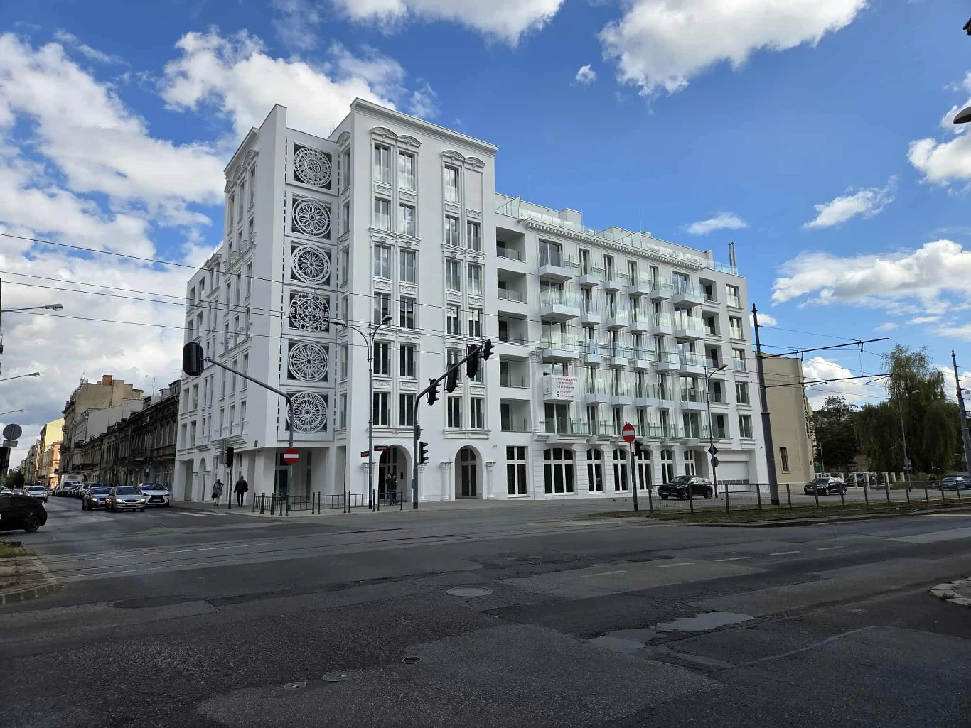 Nowoczesny, biały budynek z ozdobnymi detalami na elewacji, sześcioma kondygnacjami, dużymi oknami oraz balkonami, stojący na rogu szerokiej miejskiej ulicy z widocznymi światłami sygnalizacji.