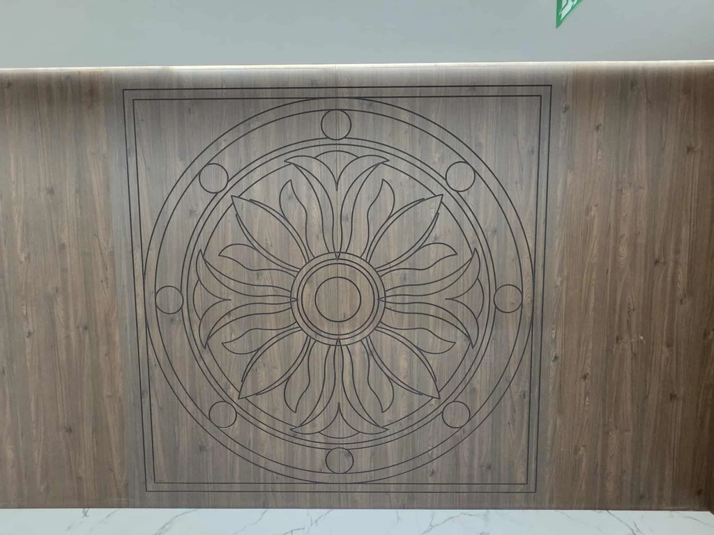 Takie motywy pojawiają się kilkukrotnie na korytarzach. Wnętrza są eleganckie Duży drewniany panel z grawerowanym wzorem geometryczno-roślinnym w centrum, utrzymany w ciepłej tonacji drewna, umieszczony nad jasnym, marmurowym podłożem.