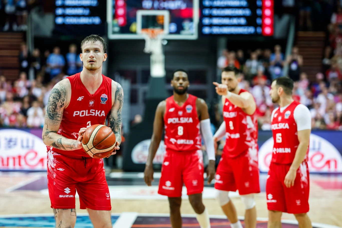 Polscy koszykarze podczas Eurobasketu 2025 Koszykarz drużyny Polski przygotowuje się do rzutu wolnego podczas meczu, otoczony przez trzech współzawodników w czerwonych strojach z napisem Polska, w tle rozmyte postacie widzów oraz tablica wyników.