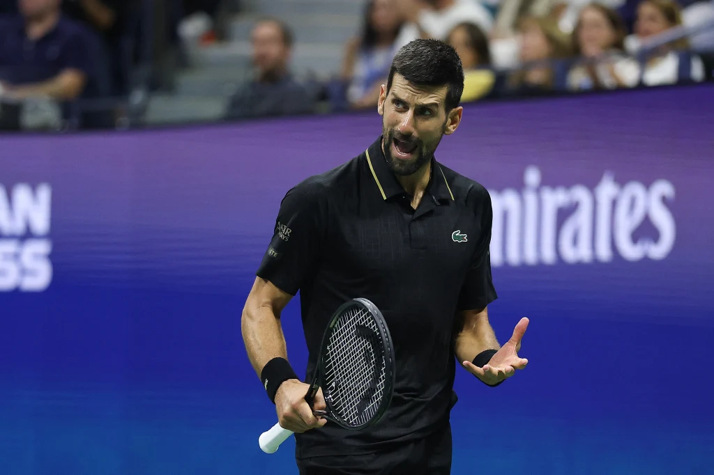 Tenisista w czarnym stroju wyraża emocje na korcie podczas meczu, trzymając rakietę tenisową w jednej ręce, w tle rozmyte sylwetki widzów i niebieskie bandy reklamowe.