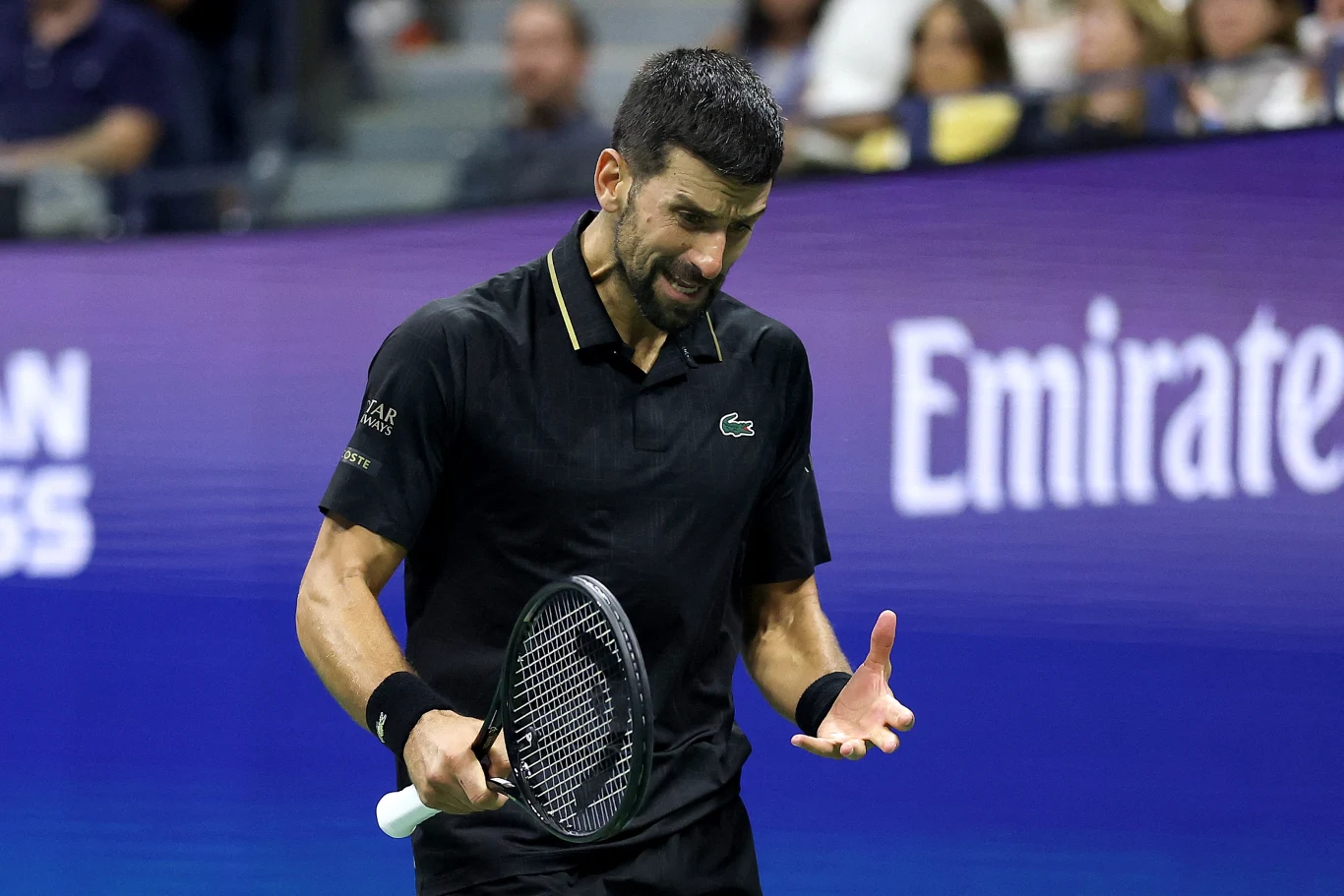 Zawodowy tenisista w czarnej koszulce podczas meczu wyraża frustrację, trzymając rakietę tenisową i gestykulując ręką, w tle widoczna jest publiczność oraz niebieska tablica z napisem Emirates.