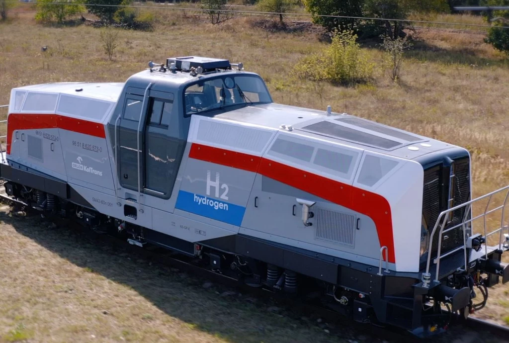 Lokomotywa wodorowa PESA SM42-6Dn. Nowoczesna lokomotywa z napędem wodorowym w srebrno-szarej kolorystyce z czerwonym pasem, stojąca na torach na tle suchej trawy oraz drzew i krzewów w oddali, na obudowie widoczny napis H2 hydrogen.