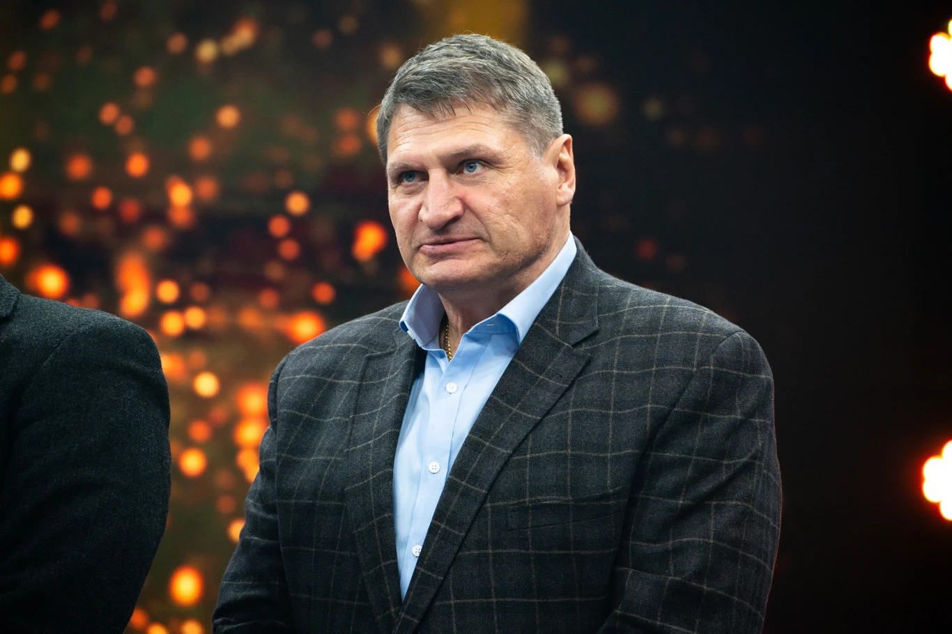 Mężczyzna w średnim wieku w eleganckiej marynarce w kratę i jasnoniebieskiej koszuli, stoi w półprofilu na tle rozmytych świateł.