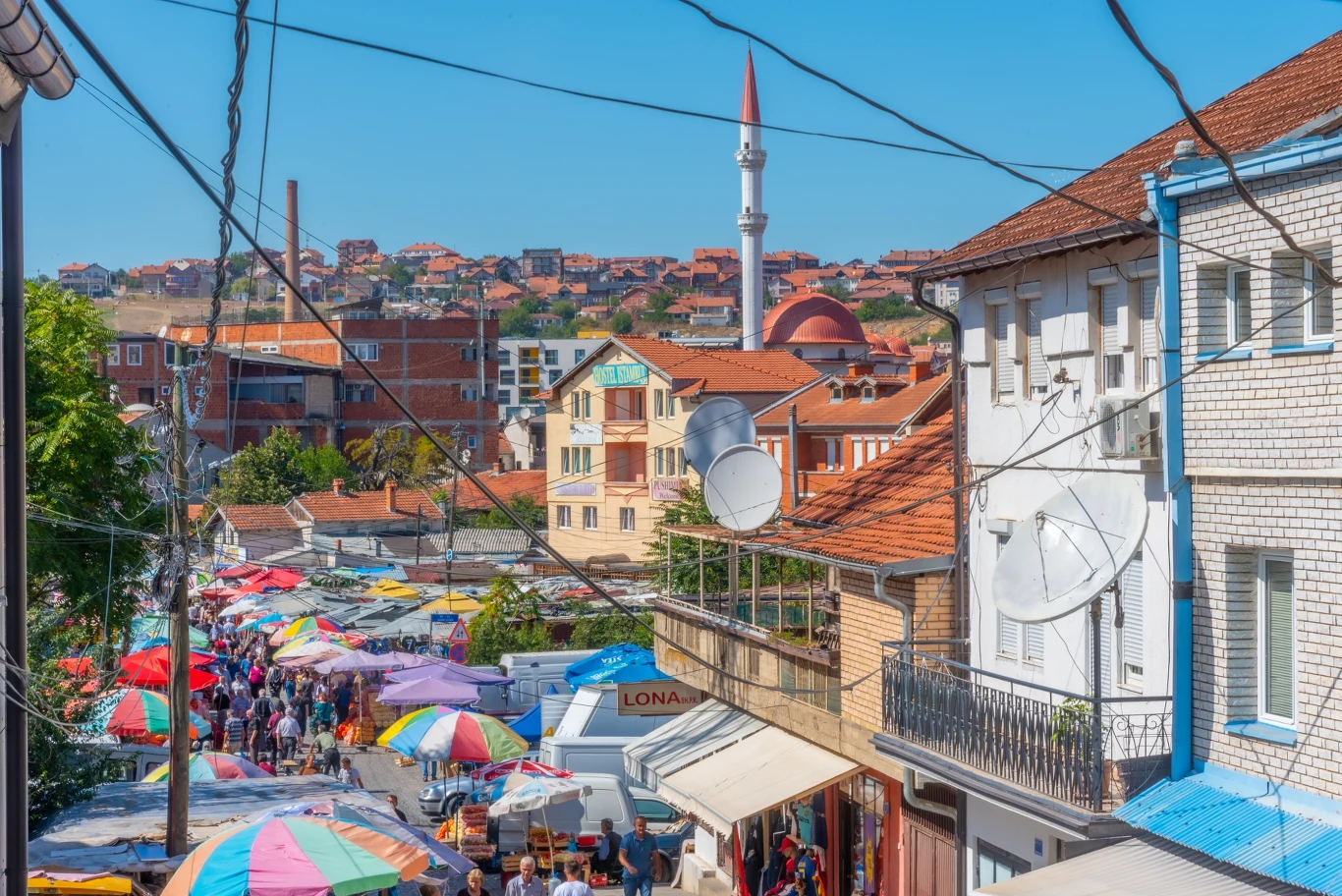 Bazar w Prisztinie Kolorowy bazar uliczny z tłumem ludzi, parasolami chroniącymi przed słońcem oraz tradycyjnymi budynkami, w tle wieża minaretu meczetu i panoramę miasta na wzgórzu.
