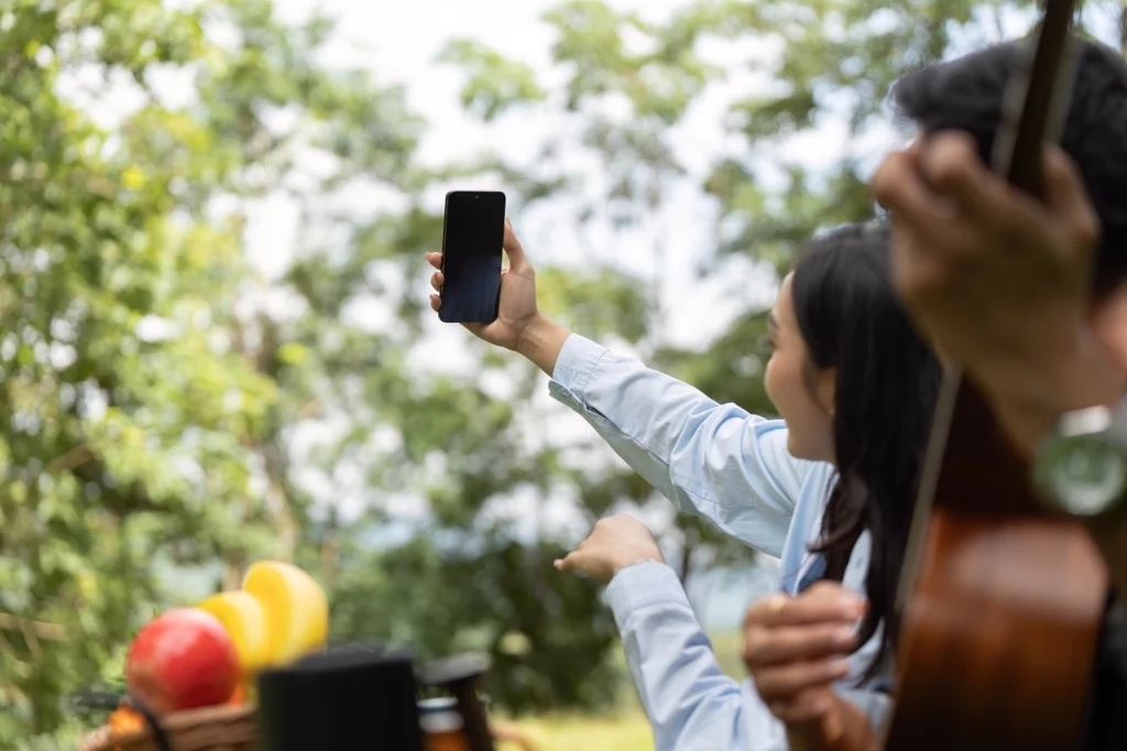 iPhone 17 Pro i Pro Max zostaną wyposażone w system trzech obiektywów Grupa osób siedzących na pikniku, kobieta robi zdjęcie telefonem, a w tle widać kosz piknikowy z owocami i osobę grającą na gitarze.