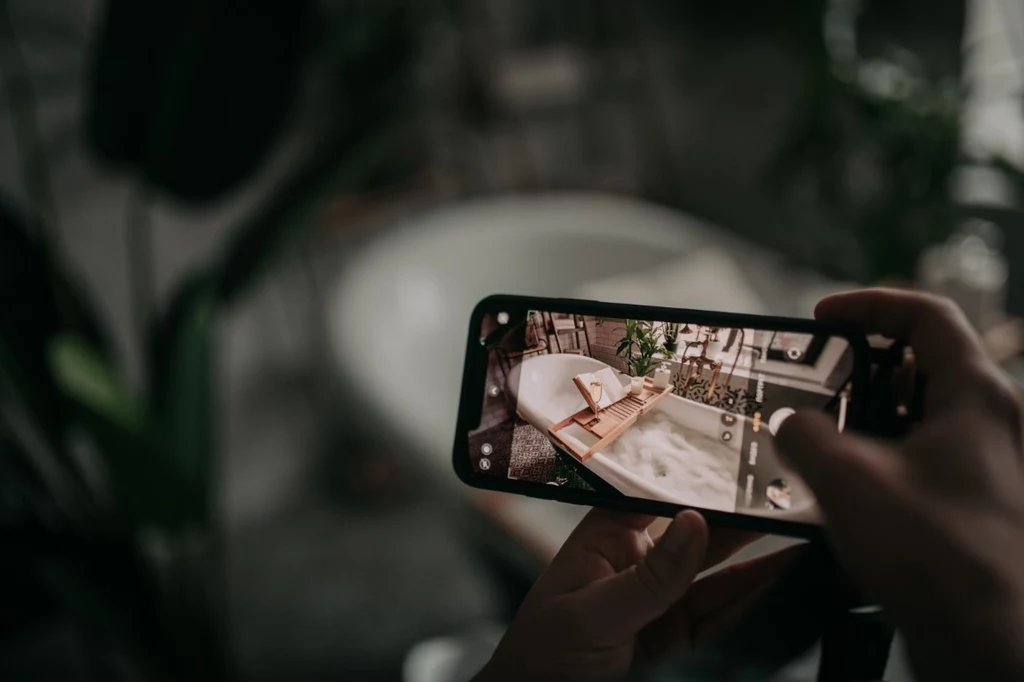 iPhone 17 z wyświetlaczem Super Retina XDR z technologią ProMotion Ręce trzymające smartfon wykonują zdjęcie stolika obok wanny z pianą. Na stoliku znajduje się książka, okulary, świeca i roślina doniczkowa, tworząc atmosferę relaksu i odpoczynku.