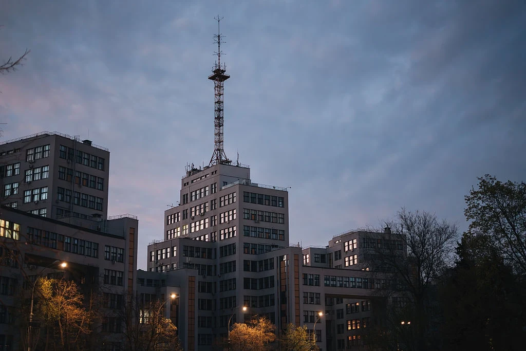 Nowoczesny budynek biurowy z anteną na dachu, otoczony drzewami, oświetlony latarniami miejskimi na tle pochmurnego nieba o zmroku.
