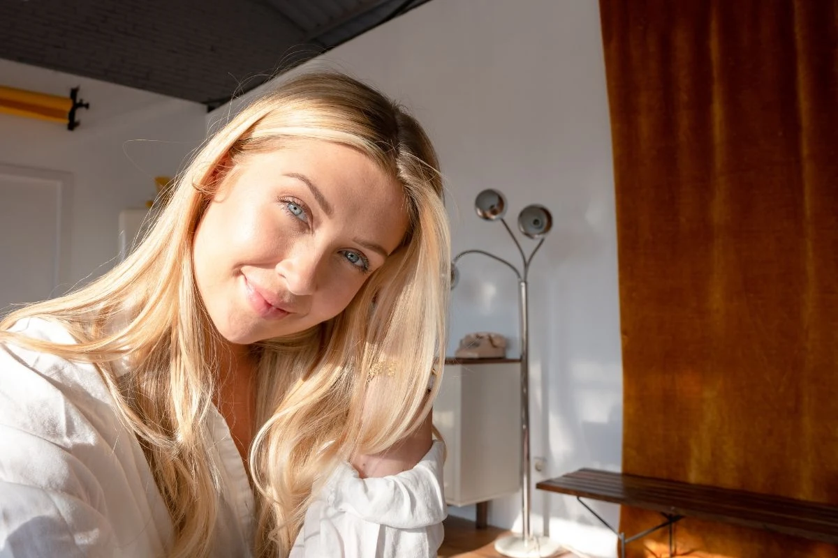 Młoda kobieta z długimi blond włosami uśmiecha się w jasnym pomieszczeniu, promienie słońca oświetlają jej twarz, w tle widoczny jest brązowy zasłon, lampa stojąca oraz minimalistyczne wyposażenie wnętrza.