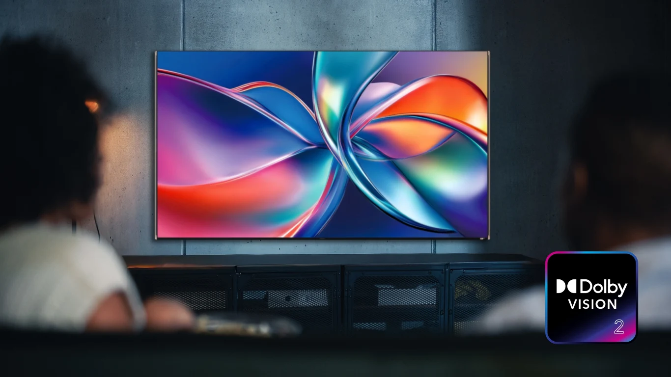 Duży telewizor z wyświetlanym kolorowym, abstrakcyjnym wzorem na ekranie, oglądany przez dwie osoby siedzące na kanapie, w prawym dolnym rogu widoczne logo Dolby Vision.