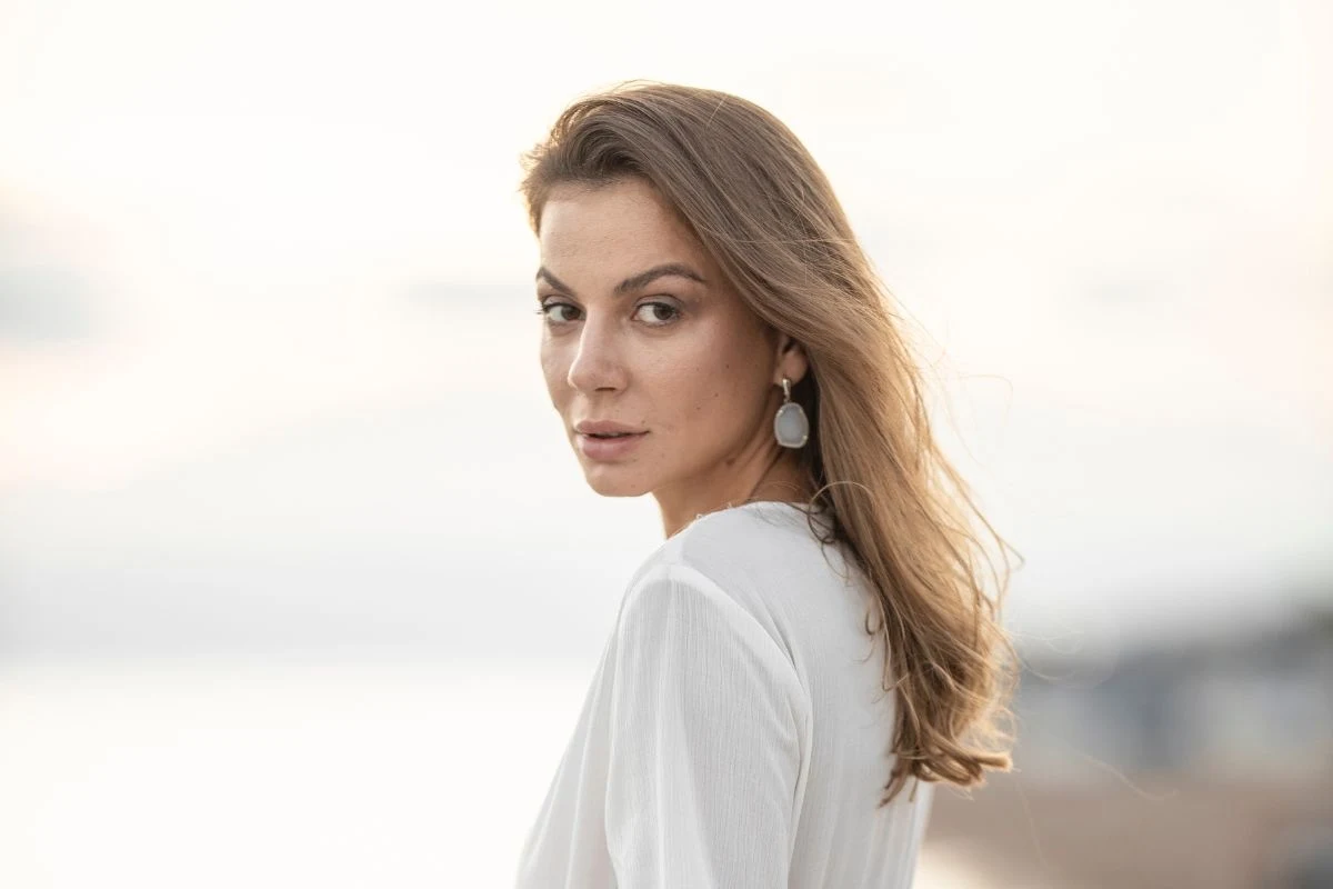 Kobieta z długimi, jasnobrązowymi włosami w białej bluzce spoglądająca przez ramię, w tle rozmyty, jasny krajobraz.