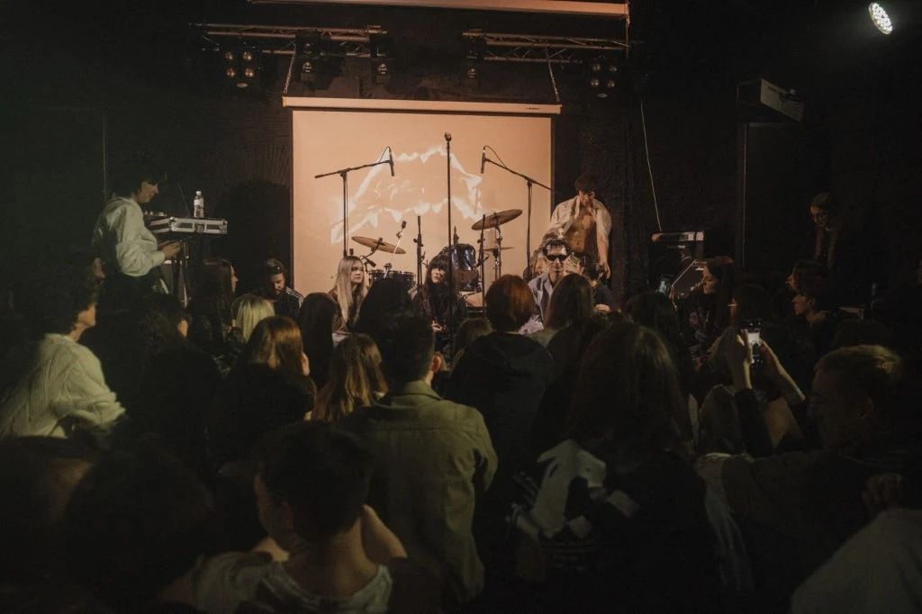 Ludzie siedzący na widowni skierowani w stronę sceny z ustawionymi instrumentami muzycznymi oraz mikrofonami, w tle jasne oświetlenie i ekran projekcyjny z grafiką.