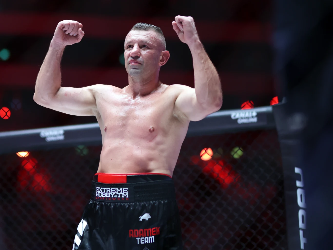 Tomasz Adamek Zawodnik sportów walki stoi w oktagonie z uniesionymi pięściami, ubrany w czarne spodenki, wyraża determinację i gotowość do walki, scena podkreślona przez czerwone światła w tle.