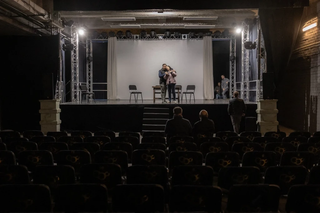 Pusta widownia teatralna, na scenie cztery osoby, z których dwie są w objęciu, a dwie kolejne znajdują się w tle; scena jest doświetlona, po bokach ustawione są krzesła, widzowie z przodu oglądają próbę lub przedstawienie.