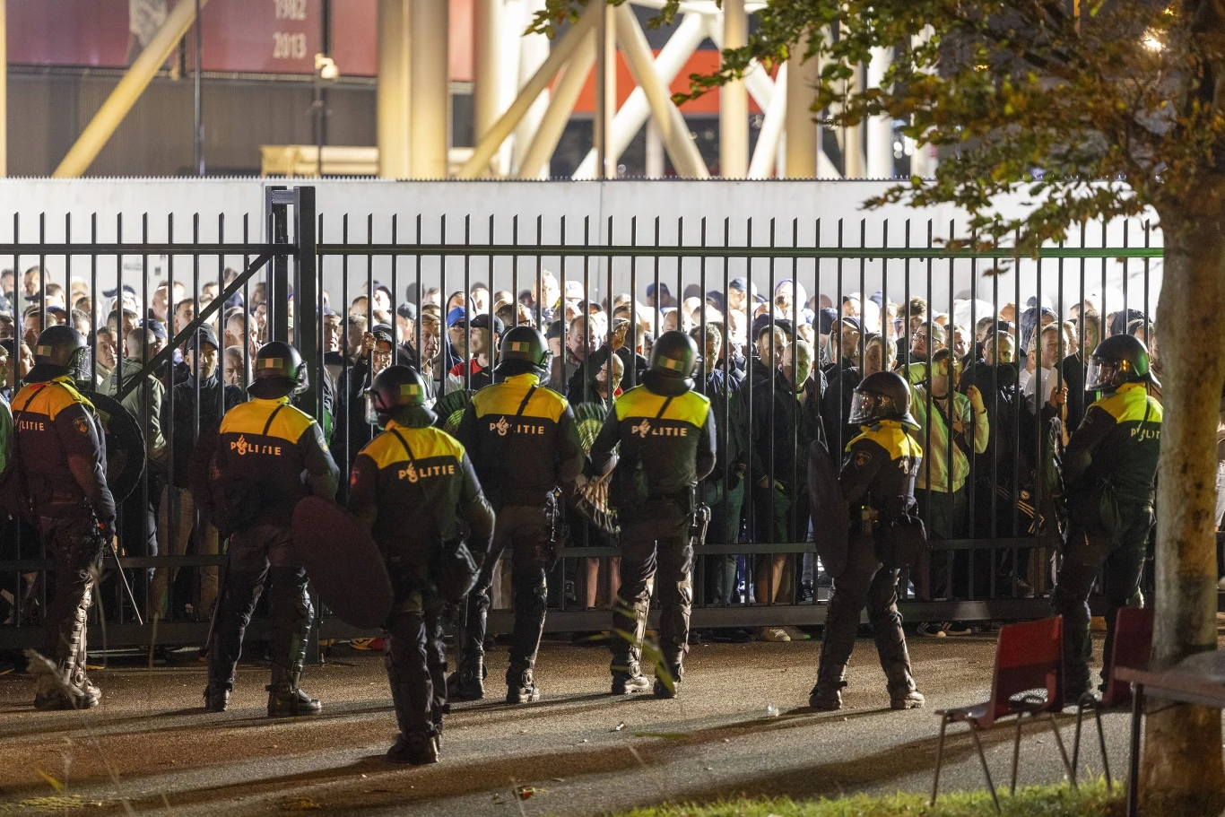Interwencja holenderskiej policji po meczu. Tak potraktowali Polaków Uzbrojeni policjanci w odblaskowych kamizelkach stoją w szeregu przy ogrodzeniu, po drugiej stronie którego zgromadził się tłum ludzi, prawdopodobnie kibiców, nocna sceneria, światło lamp ulicznych.
