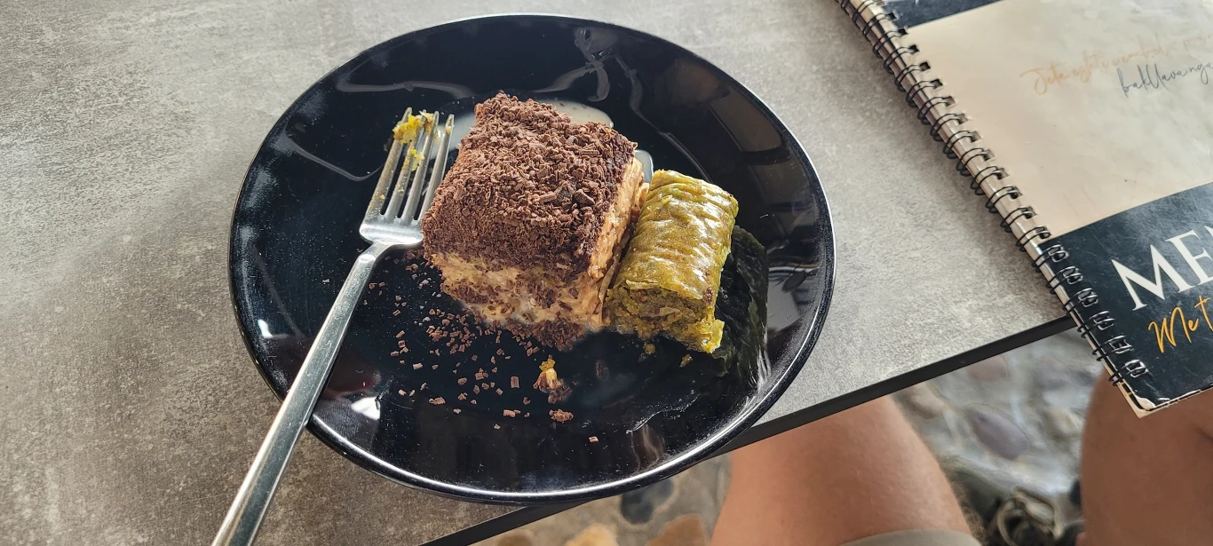Baklava to tradycyjne, bałkańskie ciasto serwowane we wszystkich kawiarniach Porcja deseru na czarnym talerzu, kawałek ciasta posypanego czekoladą obok rolady z bakaliami, widelec na talerzu, w tle menu i fragment notatnika.