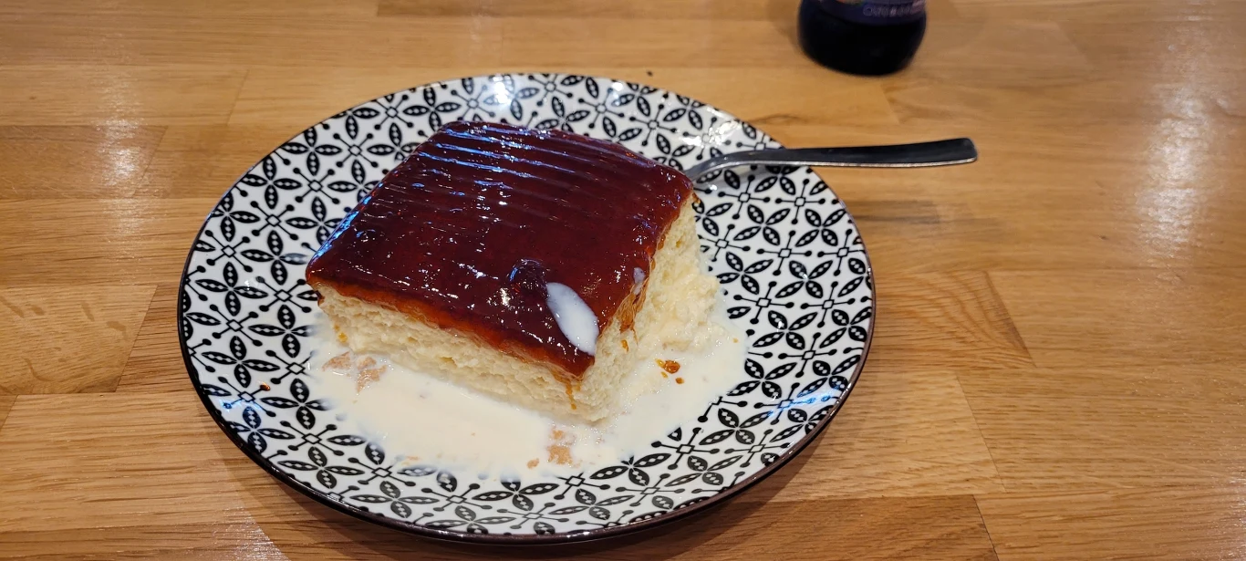 Do kawy można zamówić słynny bałkański deser - Tres Leches, czyli ciasto mleczne Kawałek biszkoptowego ciasta z galaretką na wierzchu podany na ozdobnym biało-czarnym talerzu, leżący w mlecznym sosie, z małą łyżeczką obok.