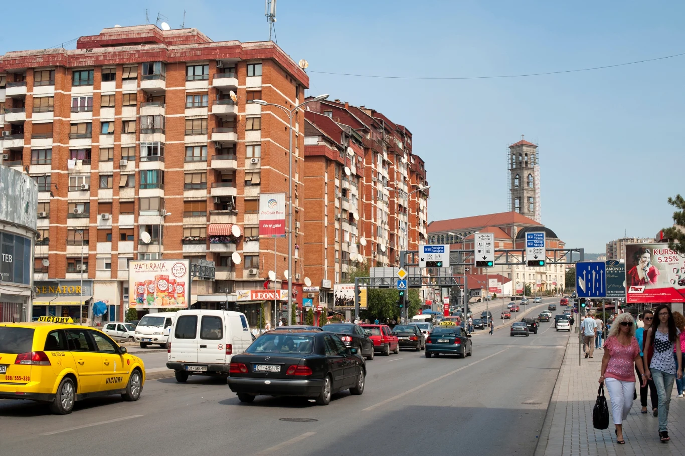 Prisztina zmaga się z chaosem na ulicach, korkami, łamaniem przepisów drogowych i brakiem parkingów Ruchliwa ulica w centrum miasta otoczona wysokimi budynkami mieszkalnymi i lokalami usługowymi, liczne samochody i taksówki na jezdni, przechodnie spacerują chodnikiem, w tle widoczna wieża kościoła.