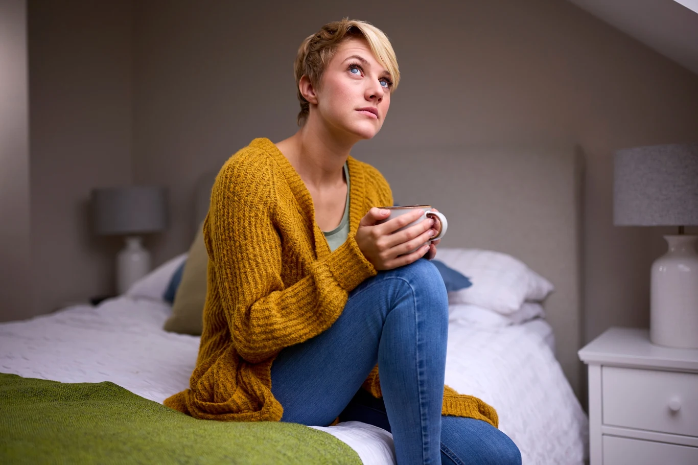 Młoda kobieta siedzi na łóżku z kubkiem w dłoniach, ubrana w żółty sweter i dżinsy, patrzy zamyślona w górę, otoczona spokojnym, ciepło urządzonym wnętrzem sypialni.