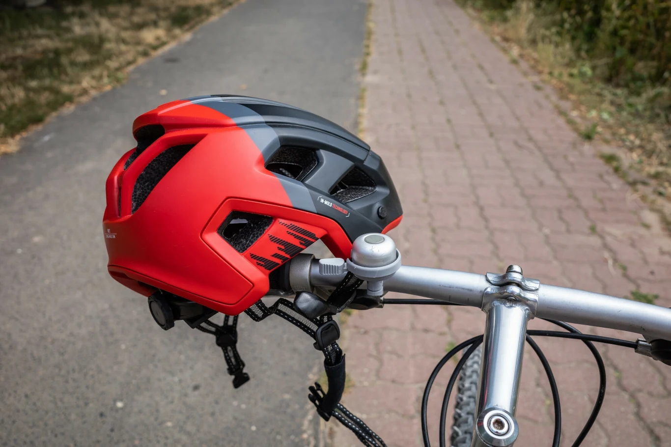 Z niezrozumiałych względów część rowerzystów jest przeciwna kaskom Czerwony kask rowerowy zawieszony na kierownicy roweru ustawionego na ścieżce rowerowej, w tle widoczna asfaltowa i brukowana nawierzchnia otoczona roślinnością.