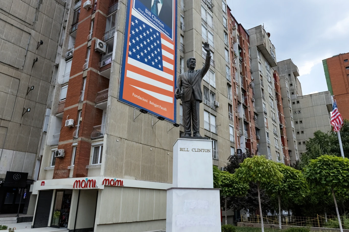 Pomnik Billa Clintona w Prisztinie pomnik Billa Clintona wzniesiony na tle wysokiego budynku mieszkalnego, obok duży billboard z portretem i flagą Stanów Zjednoczonych, w pobliżu widoczna również amerykańska flaga oraz drzewa