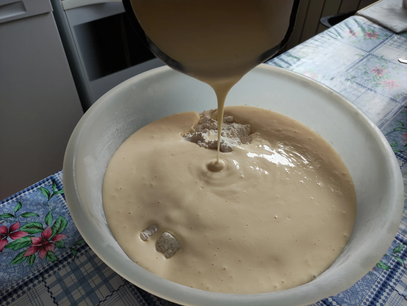 Gęsta, kremowa masa w dużej misce, do której jest właśnie dolewane nowe ciasto, leżąca na stole kuchennym z kwiecistym obrusem i naczyniami w tle.
